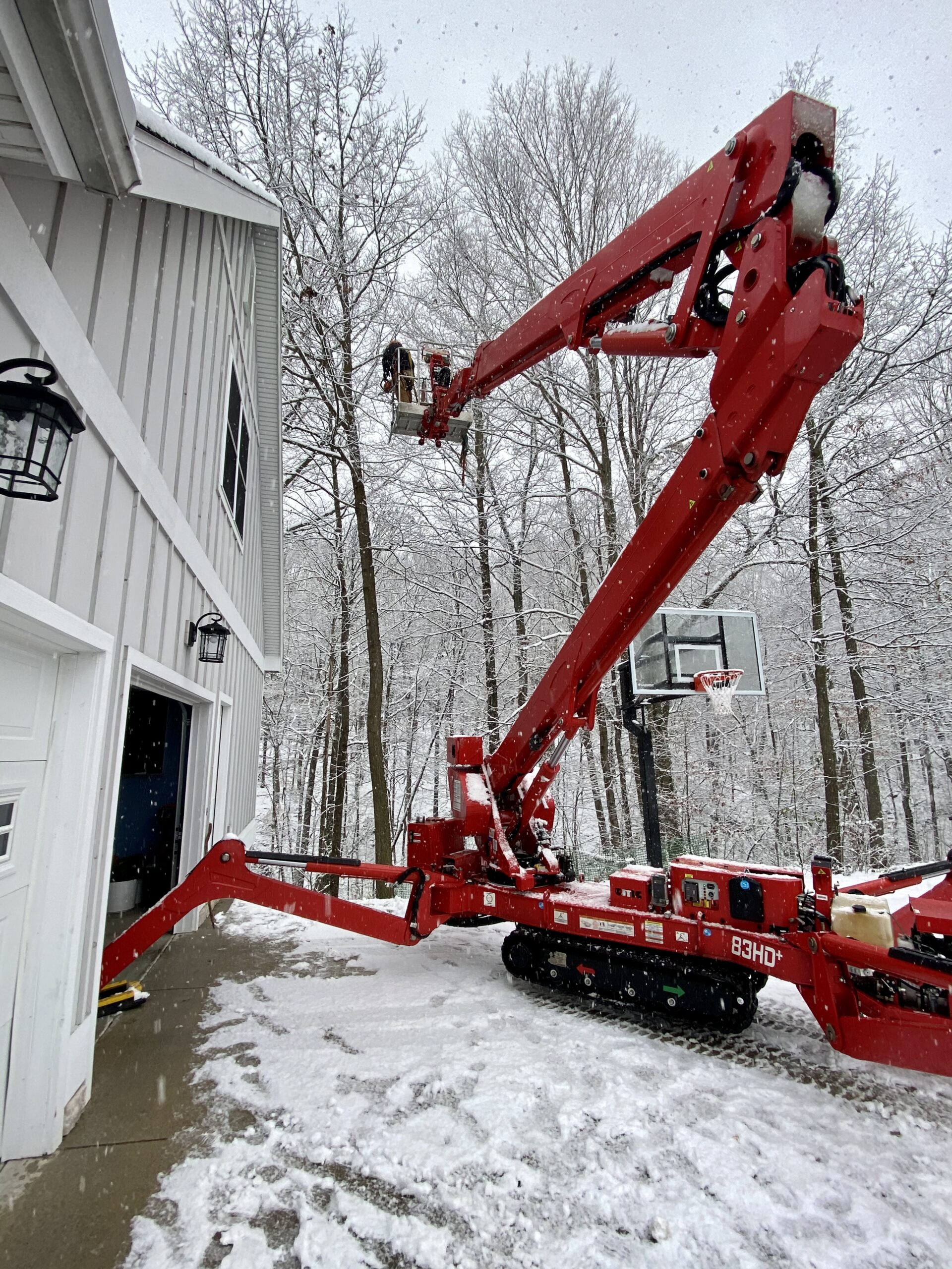 A red cherry picker extends toward a tree in a snowy landscape, with workers visible at the top